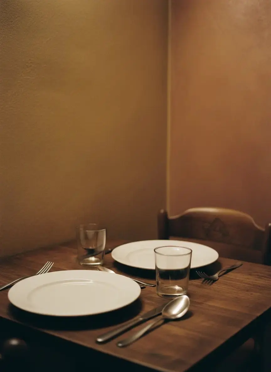 A laid table set with plain white plates and cutlery in a warm, dimly lit dining corner.