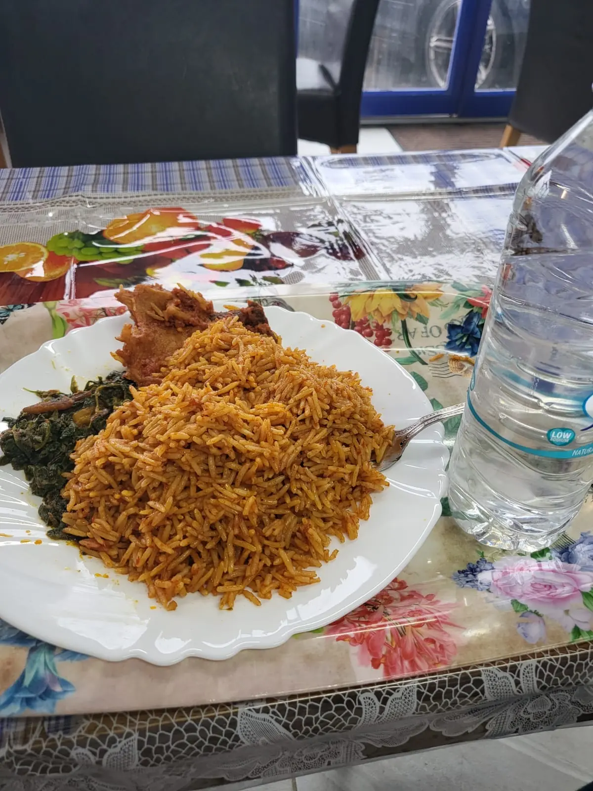 A plate of jollof rice with fried chicken and greens on a patterned table, served alongside other dishes and a bottle of water.